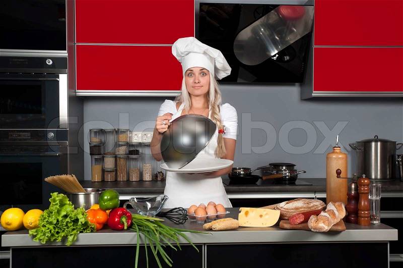Young beautiful girl chef in the kitchen with a cloche in hand, near the kitchen table with food, stock photo