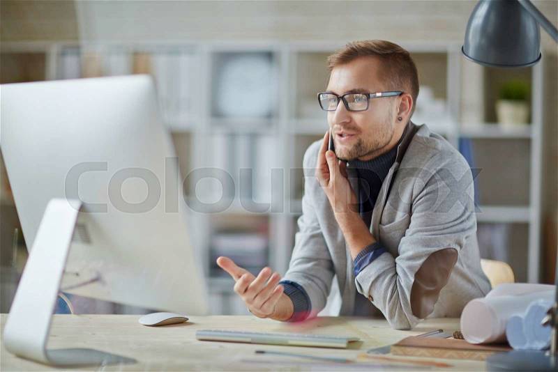 Young office worker calling in front of computer, stock photo