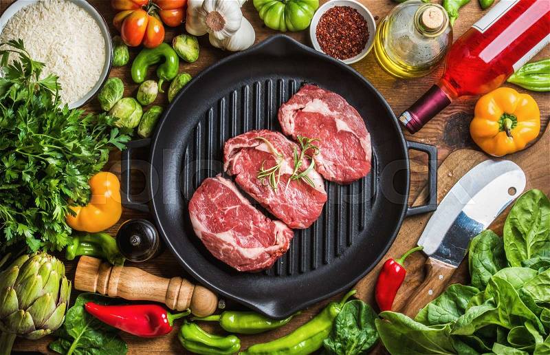 Ingredients for cooking healthy meat dinner. Raw uncooked beef steaks with vegetables, rice, herbs, spices and wine bottle over rustic wooden background, cast irom grilling pan in center. Top view, stock photo