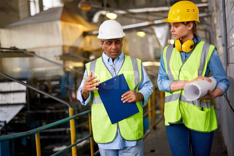 Two contemporary engineers in uniform and protective helmets discussing work, stock photo