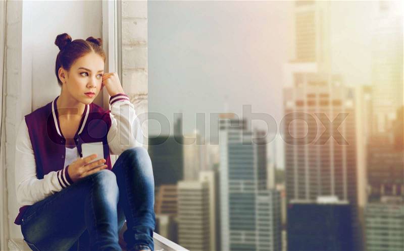 People, emotion, technology and teens concept - sad unhappy pretty teenage girl sitting on windowsill with smartphone and looking to window, stock photo