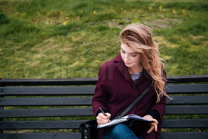 Thoughtful attractive young woman sitting on bench and writing in notebook outdoors, stock photo