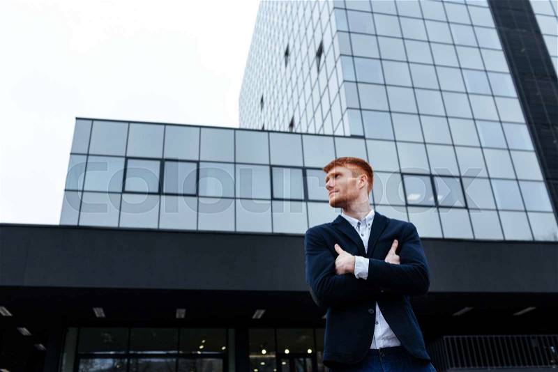 Redhead businessman standing with arms folded outdoors with glass building on background and looking away, stock photo