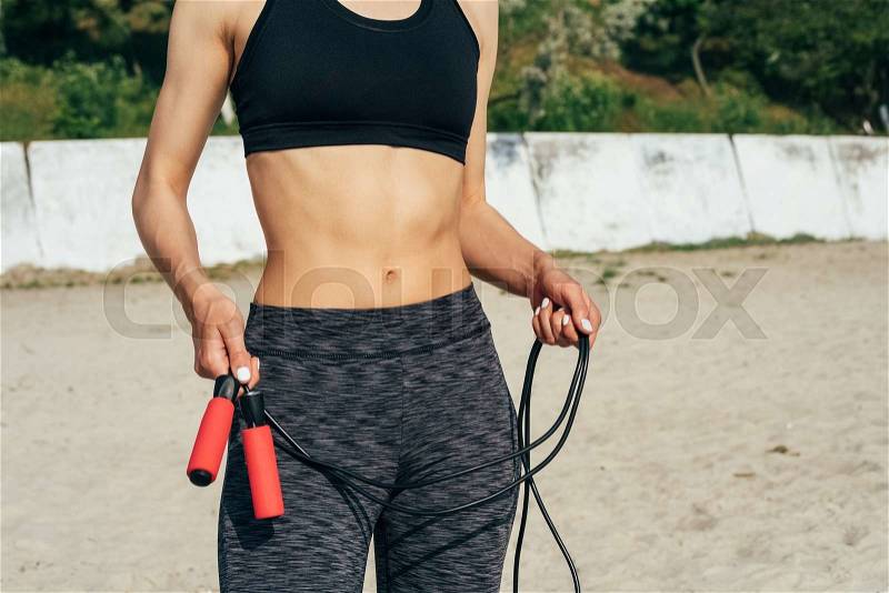 Close-up of a young athletic woman in sportswear holding a red skipping rope on the beach, stock photo