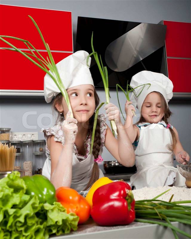 Two little girls in clothes cooks in the kitchen at the table with raw food, with bunches of green onions in the hands of, stock photo