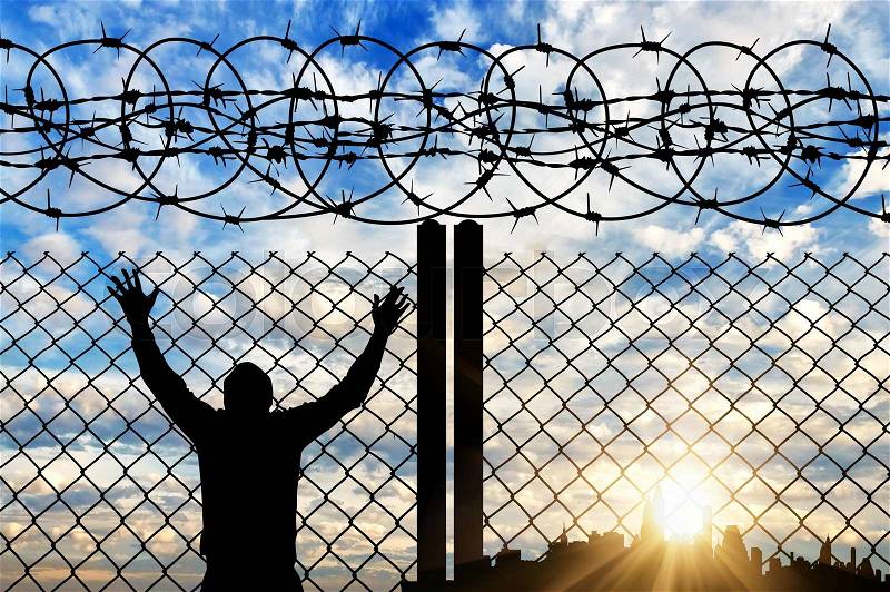 The concept of the refugees. Silhouette of a refugee near the fence with barbed wire against the evening sky and the city in the distance, stock photo