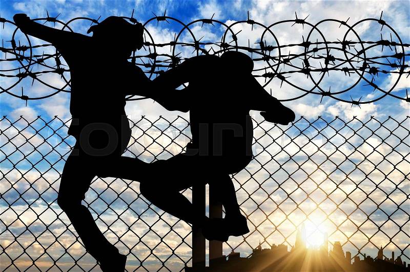 Concept of the refugees. Silhouette of refugees crossing the fence with barbed wire against the evening sky and the city in the distance, stock photo