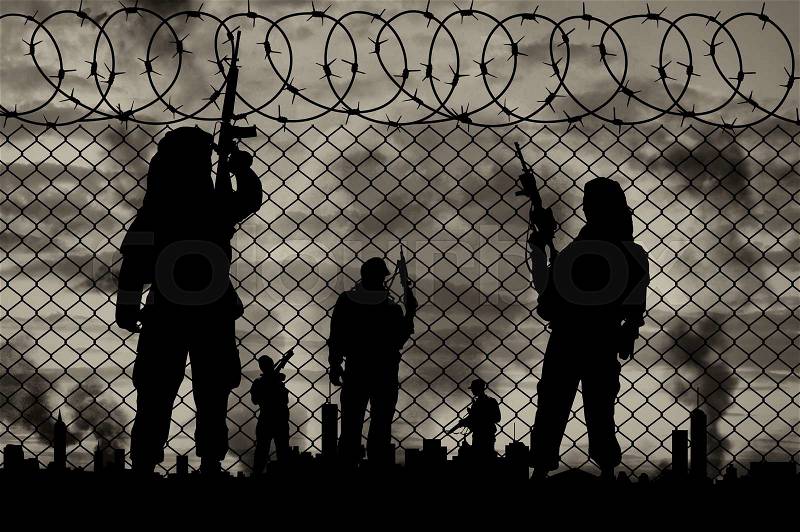 Concept of terrorism. Silhouette terrorists near the border fence in the background on the city in smoke at sunset, stock photo