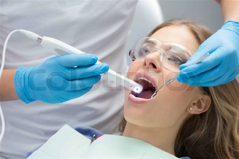 Lovely girl with opened mouth in patient bib and protective eyewear. Next to her there is a dentist in a white uniform with blue latex gloves. He diagnoses her teeth with a LED intraoral camera and a stem mouth mirror. Horizontal, stock photo