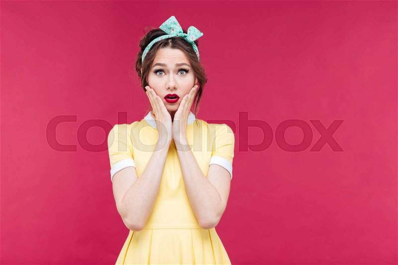 Sad shocked pretty pinup girl in yellow dress with hands on cheeks over pink background, stock photo