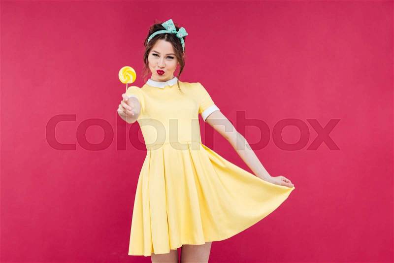 Smiling attractive pinup girl in yellow dress showing sweet lollipop, stock photo
