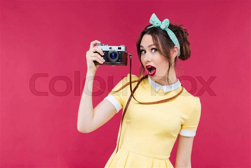 Amazed beautiful pinup girl in yellow dress holding old camera over pink background, stock photo
