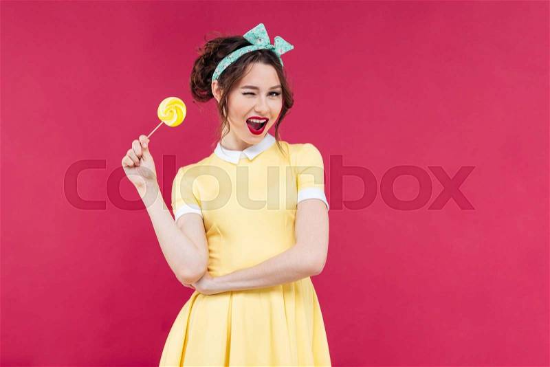 Cheerful charming pinup girl with yellow lollipop standing and winking over pink background, stock photo