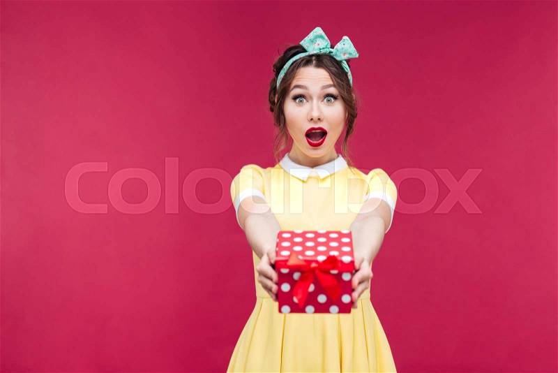 Happy surprised young woman giving you a gift box over pink background, stock photo