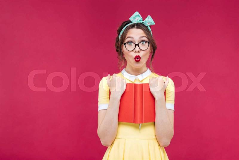 Surprised pinup girl in glasses standing and holding red book, stock photo