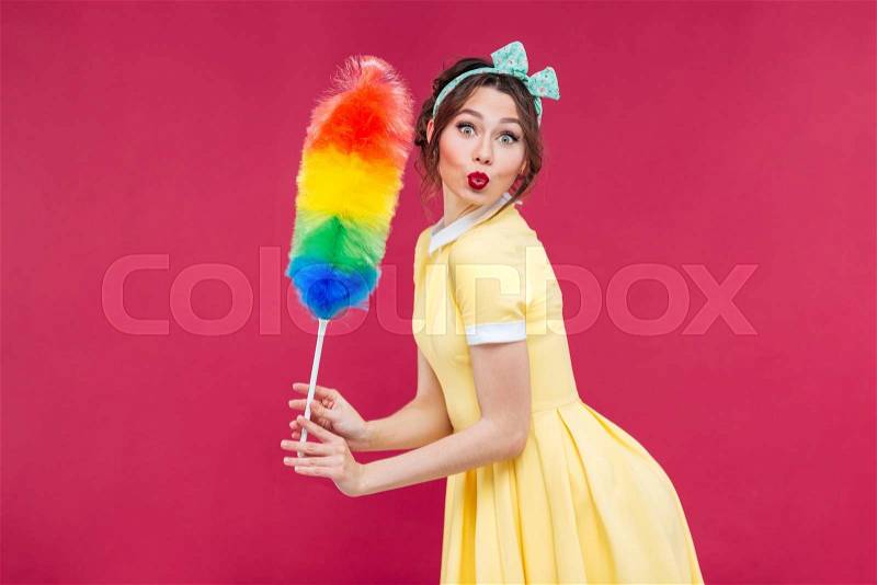 Charming playful pinup girl with colorful cleaning broom over pink background, stock photo