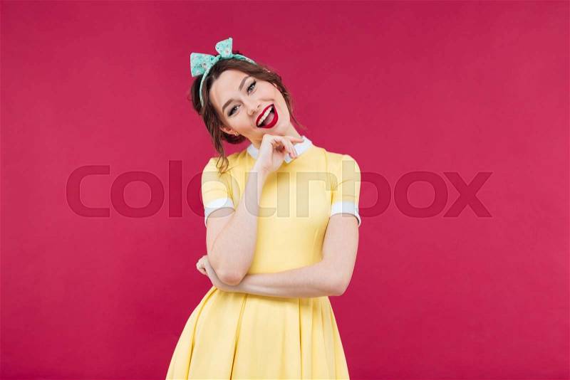 Cheerful beautiful pinup girl in yellow dress standing and smiling over pink background, stock photo