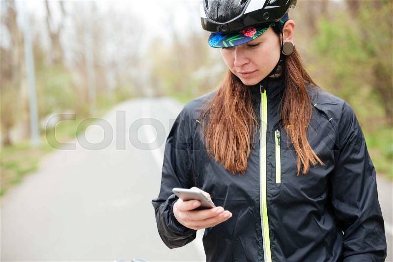 Thoughtful young woman in bicycle helmet using cell phone in park, stock photo