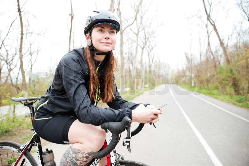 Smiling attractive young woman in bicycle helmet on bike using smartphone, stock photo