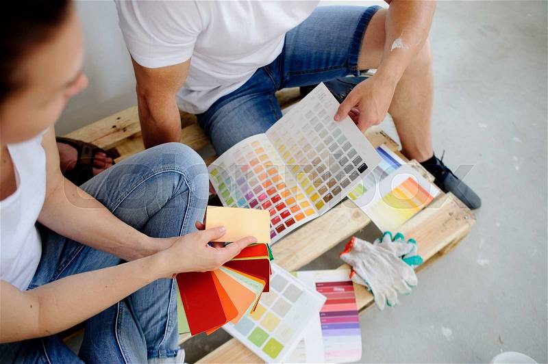 Two persons choose color of decor, sitting on the wooden pallet on a cement floor. They show each other various options. Near them various catalogs with samples are spread out, stock photo