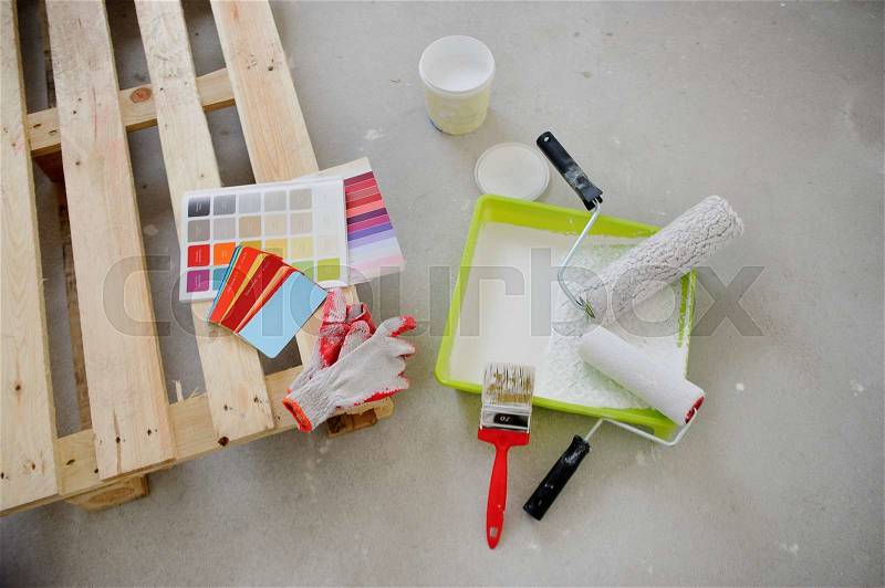 Preparation for coloring of the room.The pallet is located on a gray cement floor.Catalogs and working gloves lie on the pallet.Nearby there are a yellow tray, rollers, a brush, an open jar with paint, stock photo