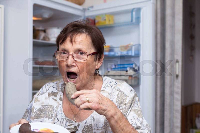 Senior woman going to eat pork liver sausage while standing in front of the open fridge in the kitchen, stock photo