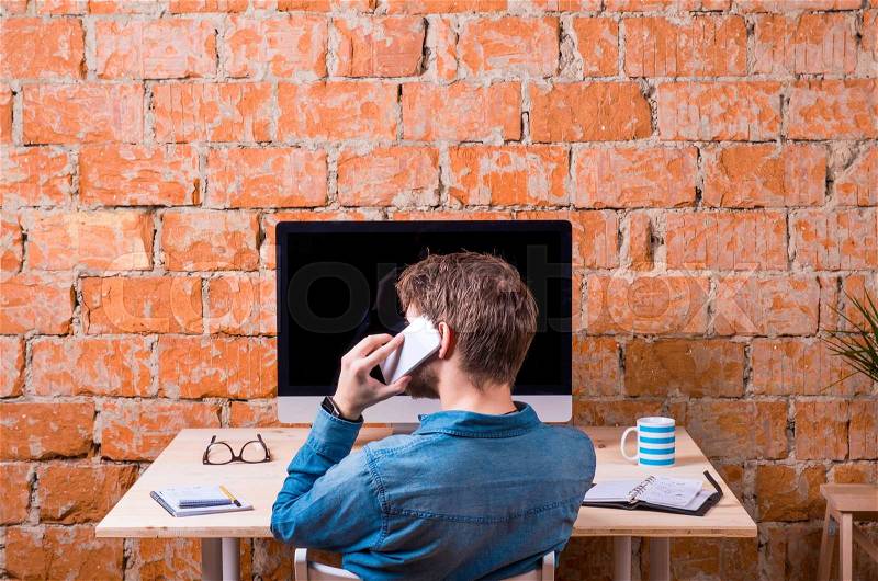 Business person sitting at office desk, talking on phone, against brick wall. Computer on the table. Coffee cup, personal organizer and various office stuff around the workplace. Rear view, stock photo