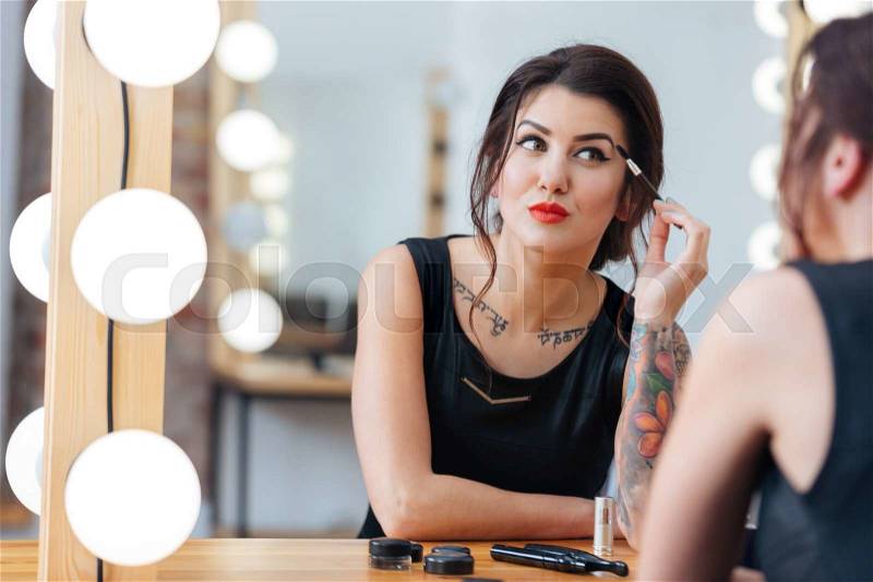 Attractive tattooed young woman sitting and doing makeup in dressing room, stock photo