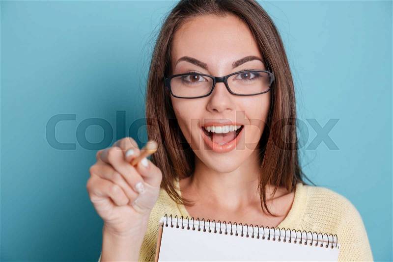 Close-up of a cute young girl pointing at camera holding notebook over blue background, stock photo