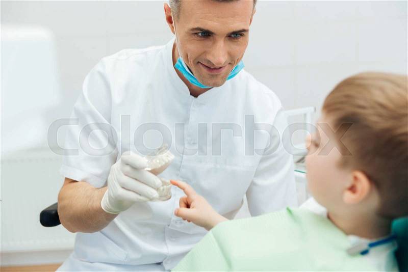 Smiling man dentist showing dental jaw model to little boy in dental clinic, stock photo