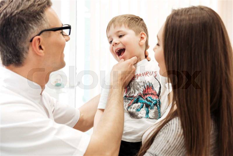 Dentist examining teeth of little boy with his mother watching at dental clinic, stock photo