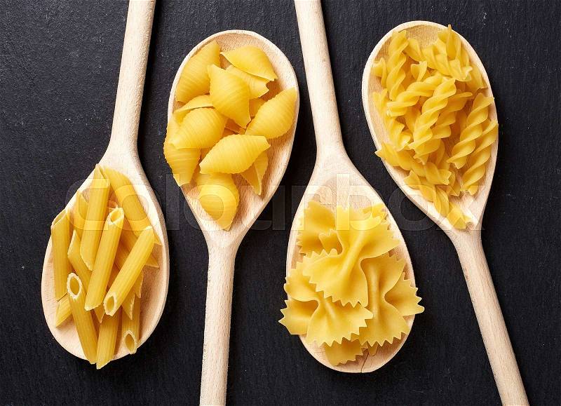 Various types of pasta in wooden spoons on black stone table, top view, stock photo
