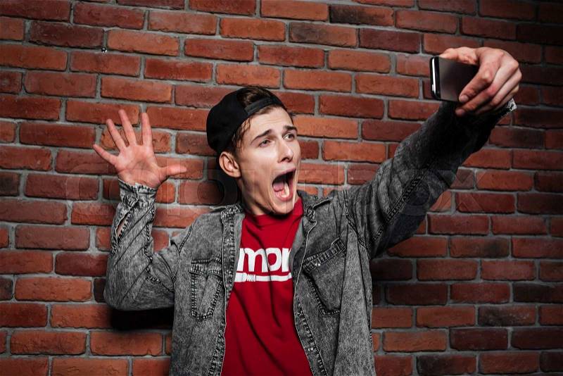 Portrait of a young man making selfie photo on smartphone over brick wall background, stock photo