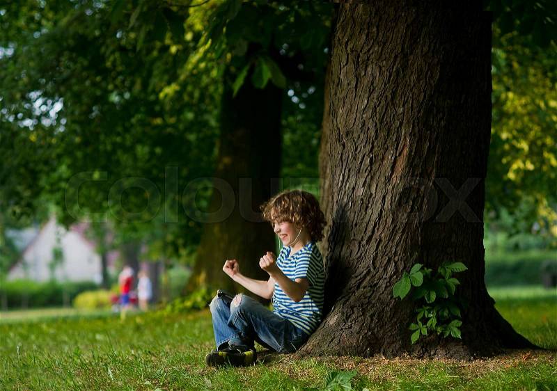 The boy with earphones in ears sits under a big tree. Something has excited him. The boy has clenched fists. On a face indignation, stock photo