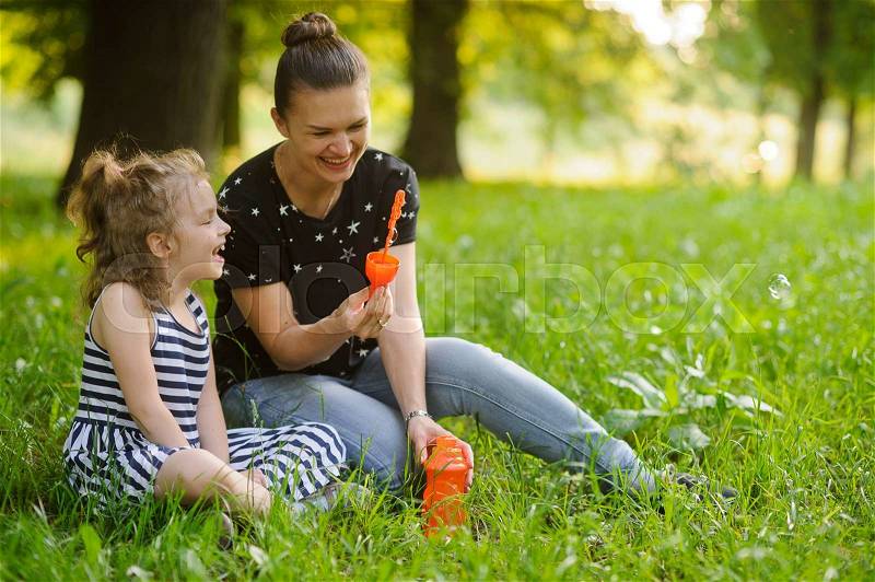 Mother with the daughter sit on a grass in park and are going to start up soap bubbles. They have a good mood and they laugh. In an environment of bright greens of park, stock photo