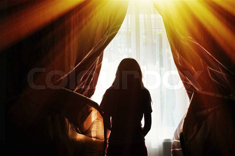 Silhouette of a young woman opens curtains at window, stock photo