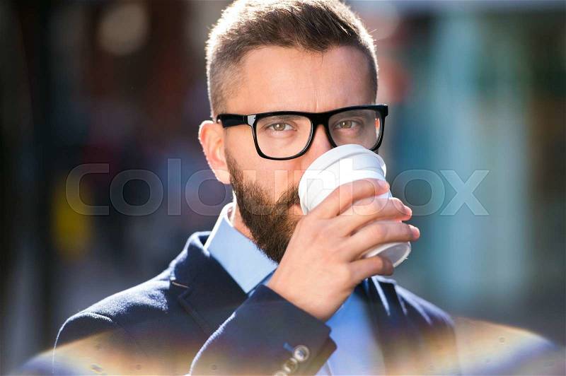 Hipster businessman holding a disposable cup and drinking coffee, walking in the street of London, wearing black eyeglasses, stock photo