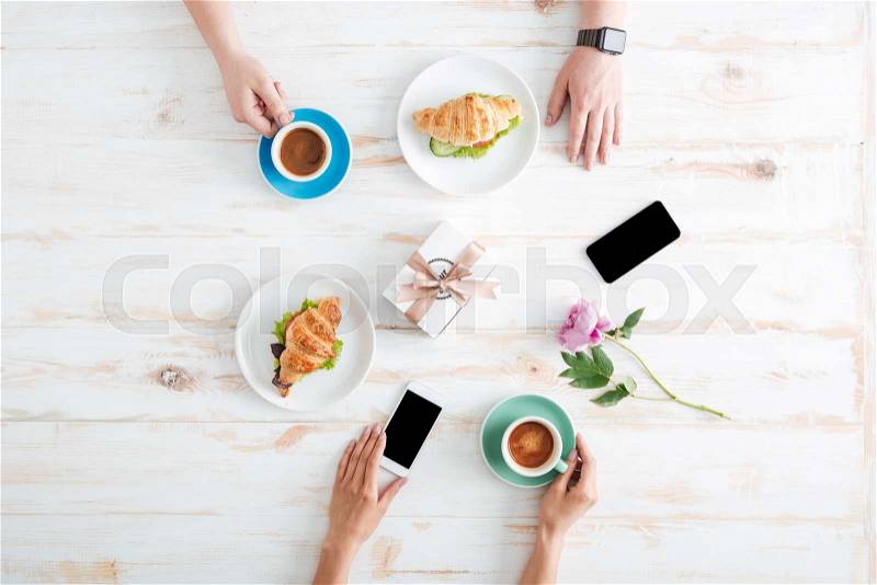 Hands of man and woman eating croissants with coffee on wooden table with blank screen cell phones, gift and flower, stock photo