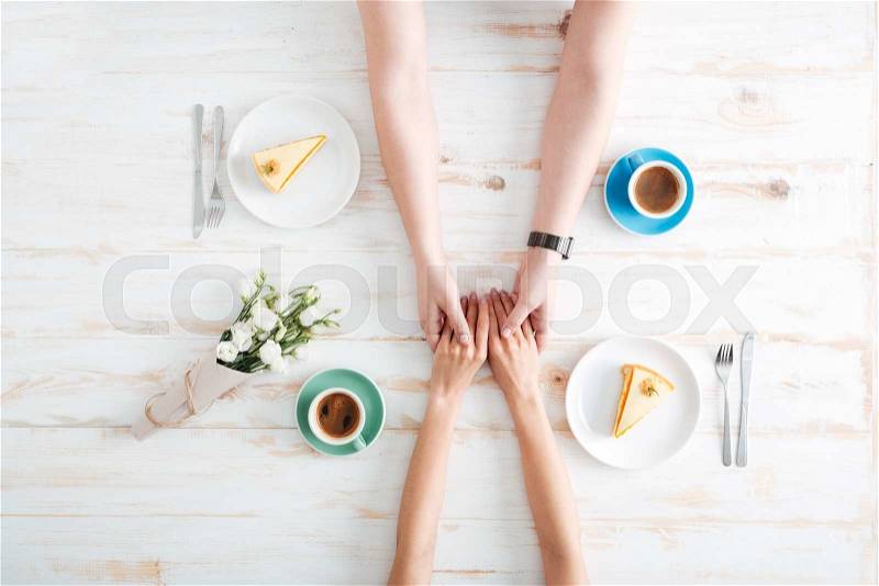 Hands of young couple holding hands and having breakfast on wooden table with coffee, cakes, gift box and flowers, stock photo