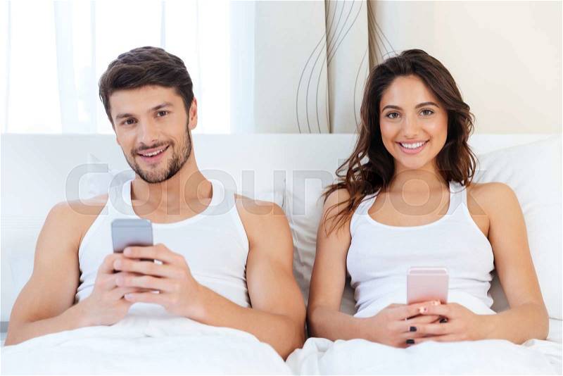 Young happy couple in a bed with mobile phones at home looking at camera, stock photo