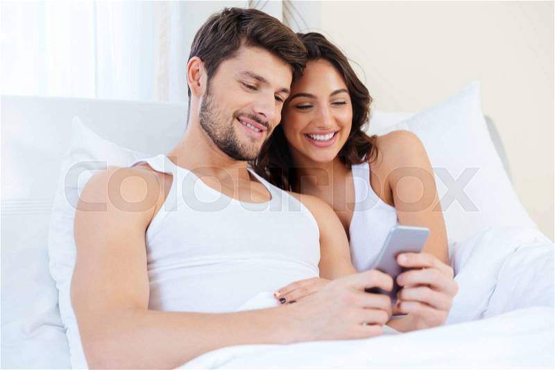 Young sweet couple in bed looking at a mobile phone at home, stock photo