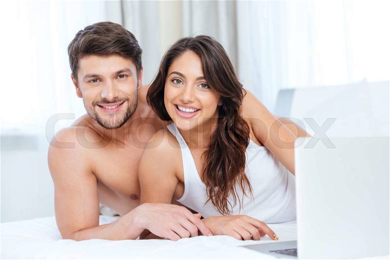 Smiling beautiful couple using laptop and looking at camera in bed at home, stock photo