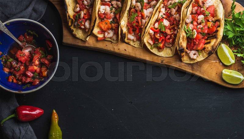 Shrimp tacos with homemade salsa, limes and parsley on wooden board over dark background. Top view, copy space. Mexican cuisine, stock photo
