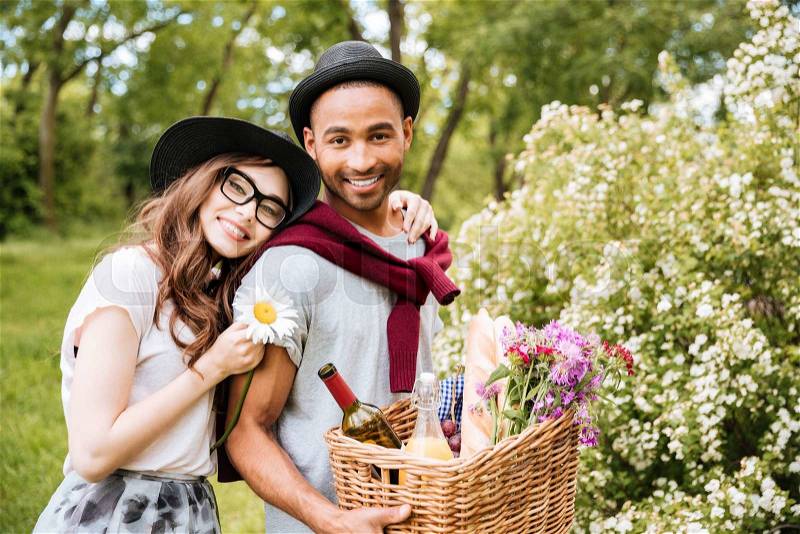 Cheeerful young couple with food and drinks for picnic standing in park, stock photo