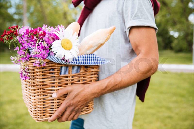 Closeup of basket with food, drinks and flowers for picnic holded by young man in park, stock photo