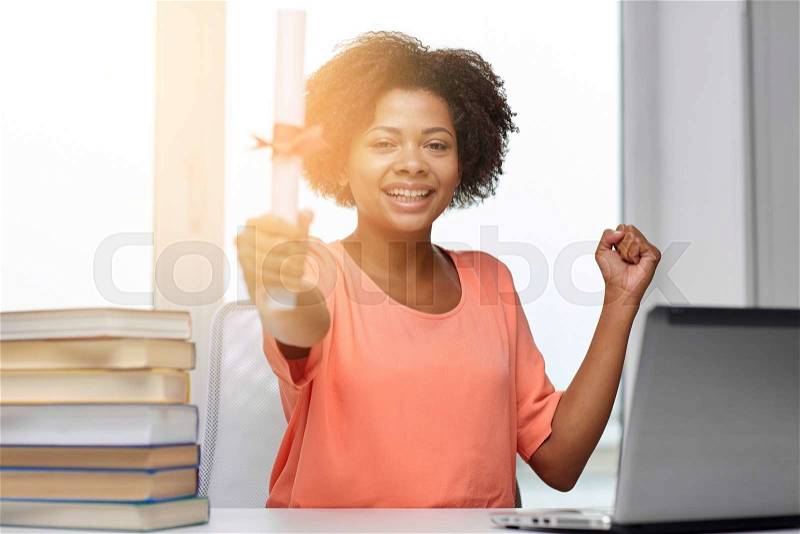 People, technology, graduation and education concept - happy african american young woman sitting at table with laptop computer, books and diploma at home, stock photo