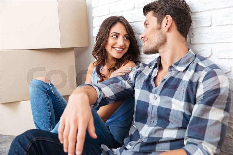 Happy couple sitting on floor around boxes after buying house and looking at each other, stock photo