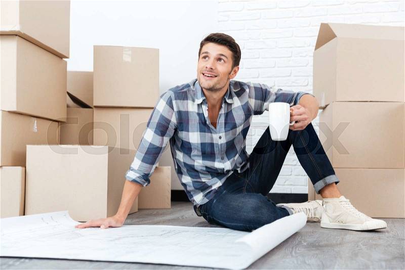 Handsome happy man looking at the house plan blueprint and holding cup indoors, stock photo
