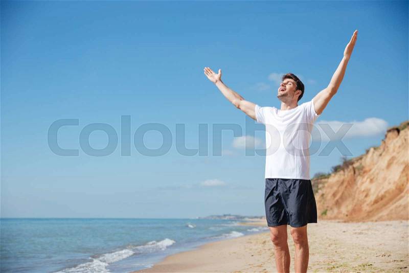 Cheerful handsome young man with outstretched arms standing on the beach, stock photo