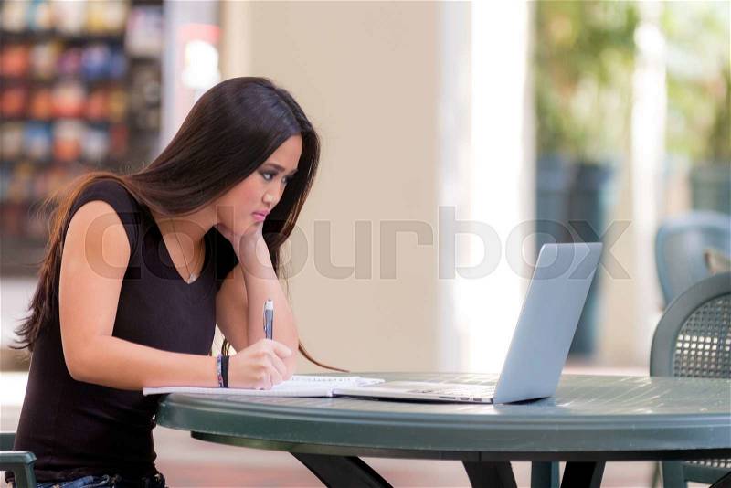 Young asian woman student sitting at table working with laptop computer and taking notes preparing for school exam, stock photo